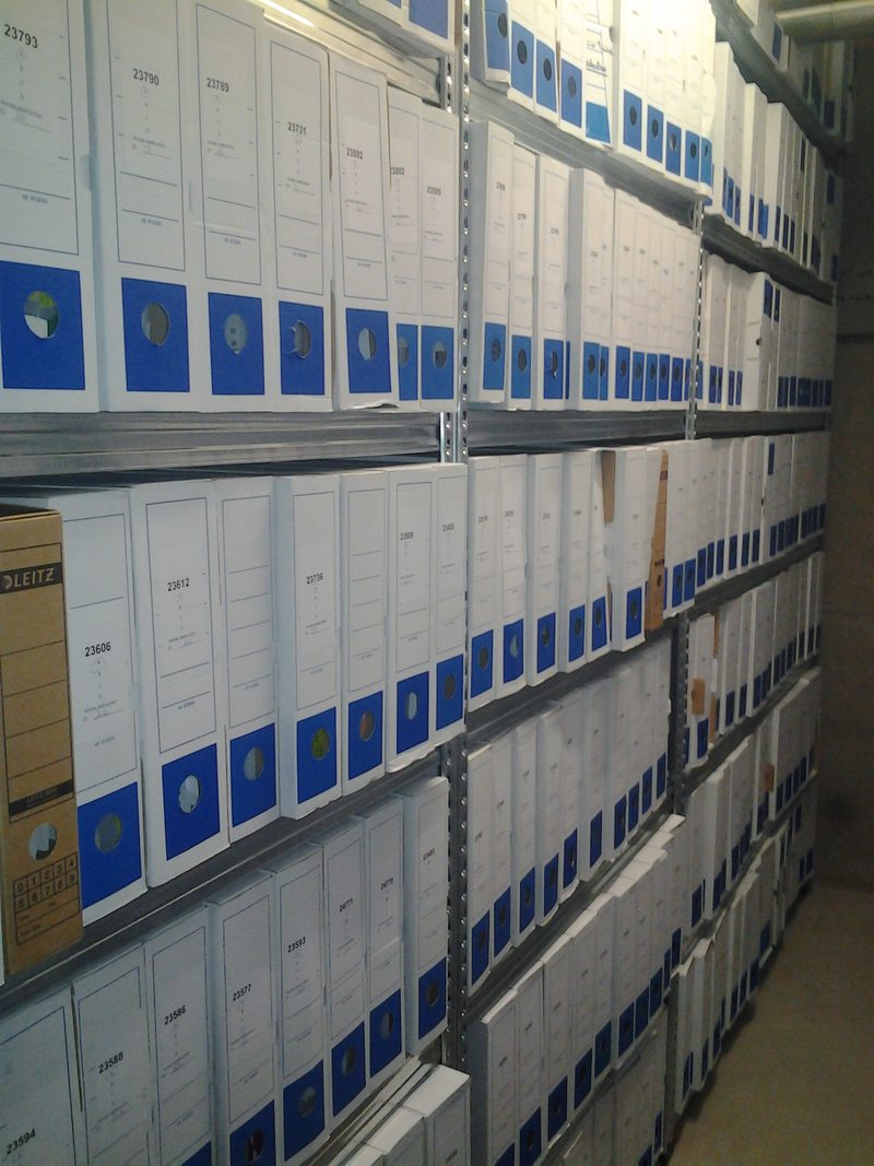 Boîtes d’archives blanches et bleues alignées sur des étagères métalliques.